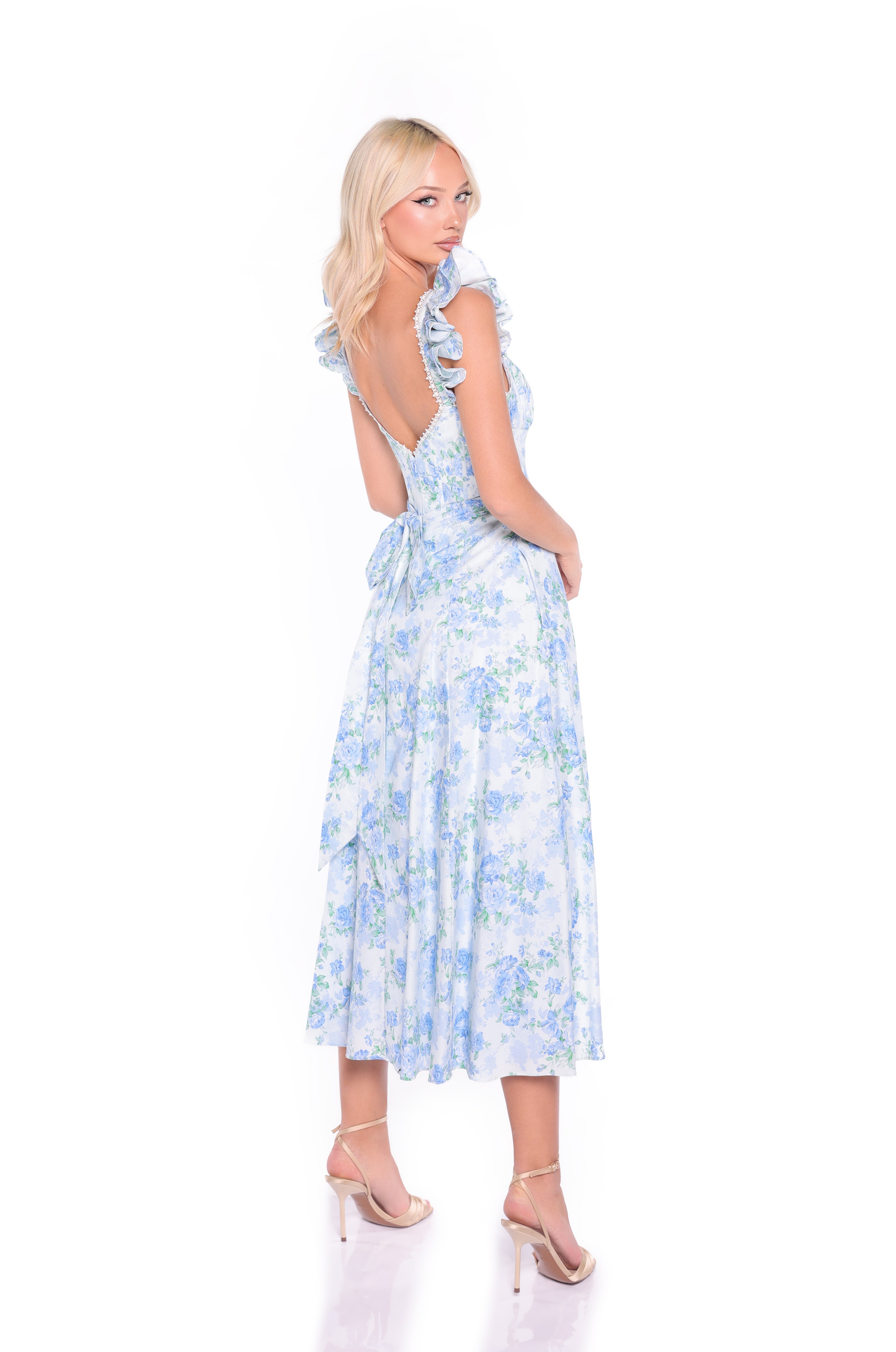 Woman wearing the Blue Floral Midi Cotton Casual Dress: Carmen, featuring ruffled straps and a back tie, stands in heels looking over her shoulder against a white background.